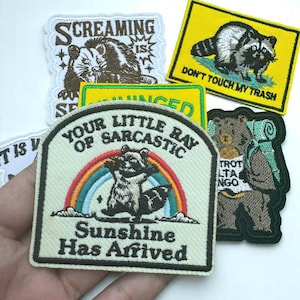 May include: A collection of embroidered patches featuring various designs. One patch shows a raccoon with the text "Your Little Ray of Sarcastic Sunshine Has Arrived." Other patches include a bear with a backpack and a raccoon with the text "Don't Touch My Trash."