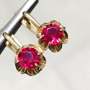 May include: A pair of gold earrings featuring round, faceted, vibrant red stones set in a scalloped, petal-like gold frame. The earrings have a hinged lever-back closure mechanism, showcasing a polished metallic finish and bright, saturated color.