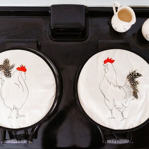 Puede incluir: Dos cubiertas redondas para la estufa con ilustraciones de gallos. Los gallos están delineados en negro con crestas rojas y detalles de plumas en blanco y negro. También se ven dos jarras pequeñas para crema y una cuchara de madera.
