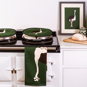 Puede incluir: Dos cubiertas de cocina verdes con un diseño de avestruz, un paño de cocina verde con un diseño de avestruz colgado de una estufa AGA blanca, una impresión enmarcada de un avestruz sobre un fondo verde, una hogaza de pan sobre una tabla de cortar de madera y una planta verde en maceta.