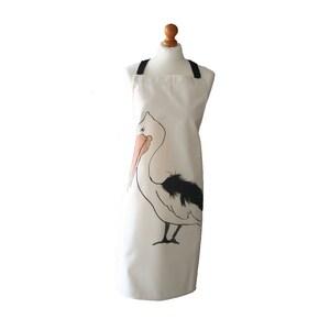 May include: A white canvas apron with black straps and a black and white illustration of a pelican with an orange beak.