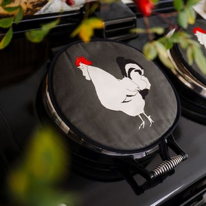 May include: Two stove top covers with a rooster design. Each cover has a white rooster with a red comb on a dark grey background. The covers are on a black stove top with a silver handle.
