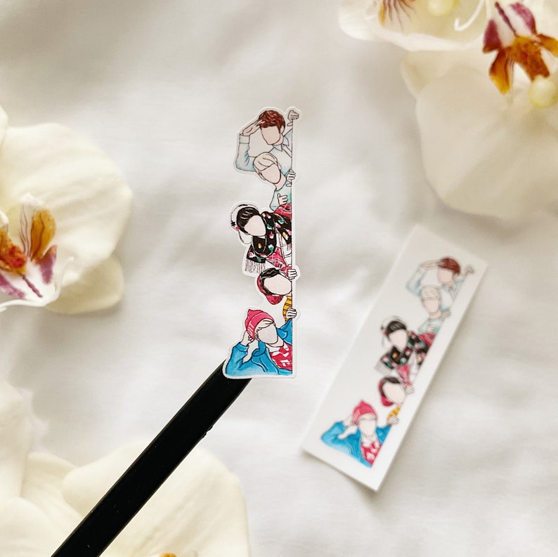 May include: A sticker featuring a cartoon illustration of several figures peeking over a white surface. The figures are dressed in various outfits, with a mix of colors including red, blue, and black. The sticker is placed on a black pencil.
