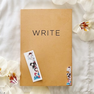 May include: A tan notebook with the word "WRITE" in black lettering. A decorative bookmark with cartoon figures is placed on the notebook. White flowers are in the background.