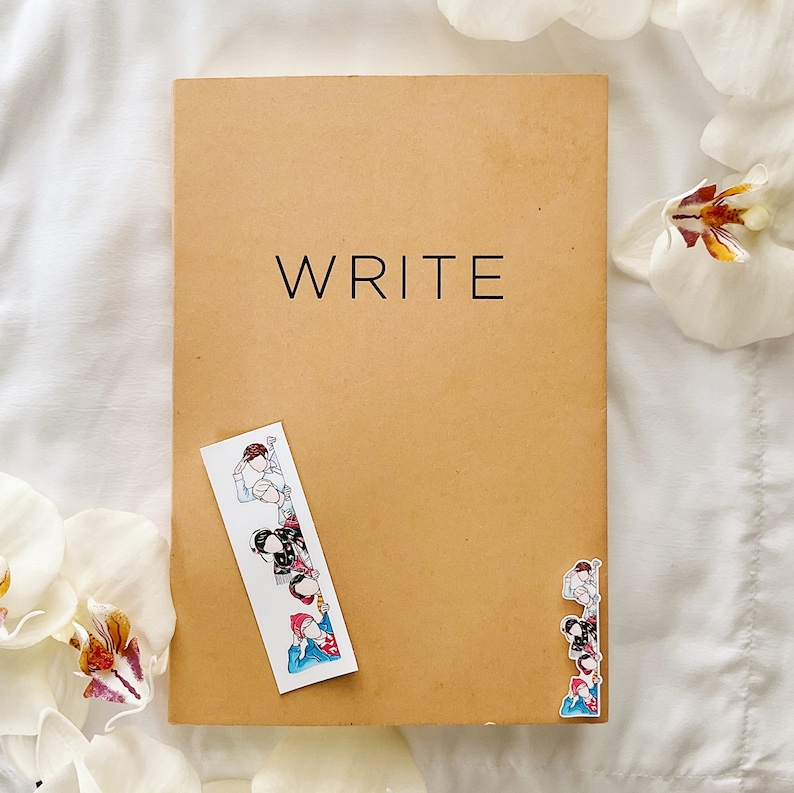 May include: A tan notebook with the word "WRITE" in black lettering. A decorative bookmark with cartoon figures is placed on the notebook. White flowers are in the background.