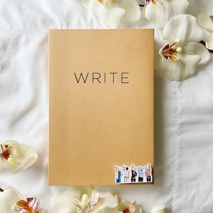 May include: A tan notebook with the word "WRITE" printed in black. A sticker depicting a group of people with raised hands is on the bottom right corner. White flowers surround the notebook on a white background.