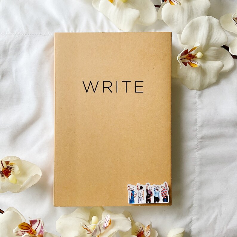 May include: A tan notebook with the word "WRITE" printed in black. A sticker depicting a group of people with raised hands is on the bottom right corner. White flowers surround the notebook on a white background.