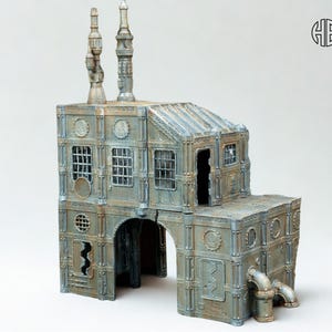 May include: A detailed miniature industrial building with a weathered, metallic finish. The model features multiple levels, arched entryways, windows, and smokestacks. The building has a distressed, aged appearance, with pipes and other industrial details.