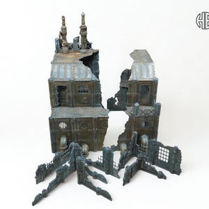 May include: A miniature, weathered, industrial building model, split into sections. The model features a distressed, metallic finish with blue and brown tones, and includes architectural details like chimneys and windows. The pieces are arranged on a white background.