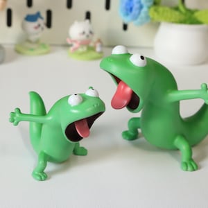 Könnte beinhalten: Zwei grüne Eidechsenfiguren mit offenem Mund und rosa Zungen. Die Eidechsen haben weiße Augen und sind mit ausgestreckten Armen posiert. Die Figuren stehen auf einer weißen Oberfläche, mit anderen kleinen Figuren im Hintergrund.