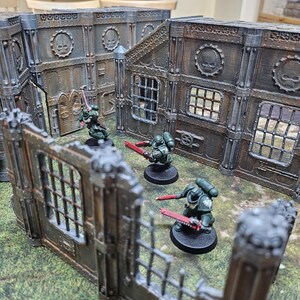 May include: A tabletop wargaming scene featuring miniature figures and terrain. The figures are painted in dark green armor and wield red weapons. The terrain includes modular, weathered-looking walls with skull emblems and window details, creating a battleground setting.