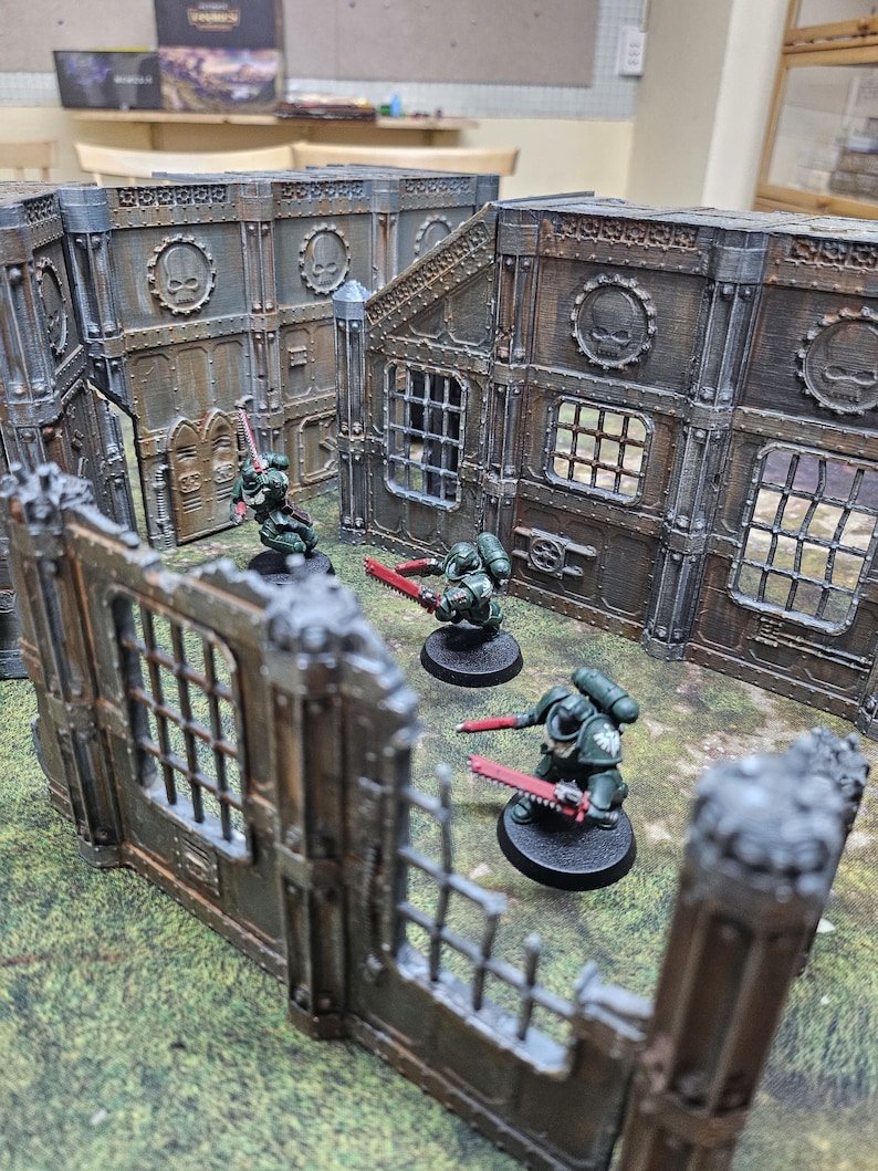 May include: A tabletop wargaming scene featuring miniature figures and terrain. The figures are painted in dark green armor and wield red weapons. The terrain includes modular, weathered-looking walls with skull emblems and window details, creating a battleground setting.
