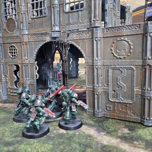 May include: Miniature figurines of green-armored soldiers with red weapons are positioned in front of a detailed, weathered industrial building. The building has arched doorways and a skull emblem. The scene is set on a green, textured surface.