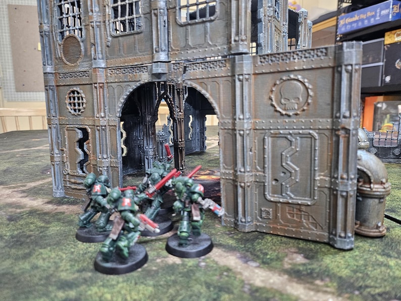 May include: Miniature figurines of green-armored soldiers with red weapons are positioned in front of a detailed, weathered industrial building. The building has arched doorways and a skull emblem. The scene is set on a green, textured surface.