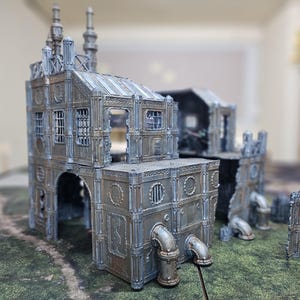 May include: A detailed miniature model of a futuristic industrial building, featuring multiple levels, arched doorways, and intricate pipework. The model is primarily gray with metallic accents, suggesting a weathered or aged appearance. Windows and architectural details are visible.