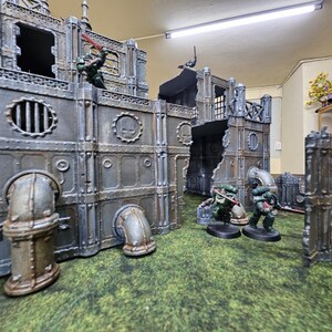 May include: A miniature war game scene featuring a detailed, weathered industrial cityscape with figures in combat. The structures are gray with metallic accents, and the figures are in green armor. The terrain is a green, textured surface.