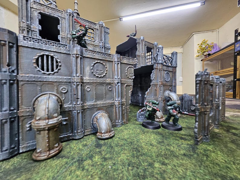 May include: A miniature war game scene featuring a detailed, weathered industrial cityscape with figures in combat. The structures are gray with metallic accents, and the figures are in green armor. The terrain is a green, textured surface.