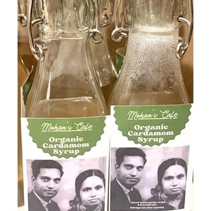 Peut inclure: Deux bouteilles en verre transparent de sirop de cardamome biologique Mohan's Cafe avec des bouchons rouges. Chaque bouteille a une étiquette avec une photo vintage d'un couple et les ingrédients.