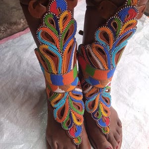 May include: Handmade leather sandals with vibrant beaded embellishments. The sandals feature a brown leather base with colourful beadwork in shades of blue, orange, green, and yellow. The design includes decorative straps and a toe post.