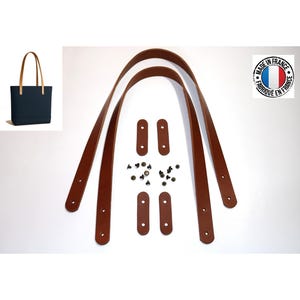 Könnte beinhalten: Zwei lange braune Lederriemen für Taschen mit vier kleinen rechteckigen Lederverstärkungen und einem Set aus Metallnieten. Ein kleines Bild zeigt eine dunkelblaue Tragetasche mit diesen braunen Lederriemen. Enthält ein kreisförmiges Logo.