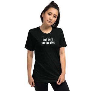 Puede incluir: Camiseta negra con el texto blanco "just here for the plot". La camiseta tiene cuello redondo y parece estar hecha de un material suave. La modelo lleva pantalones negros.