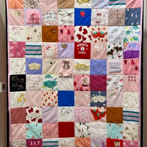 May include: A patchwork quilt made from various baby clothes, featuring a variety of colors and patterns. The quilt includes squares with strawberries, hearts, and the text "Manchester Football". The quilt is hanging on a wall.
