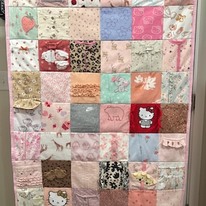 May include: A memory quilt constructed from various baby clothing pieces, featuring a patchwork design with diverse patterns including Hello Kitty, strawberries, giraffes, and floral prints. The quilt is bordered with a light pink fabric trim.