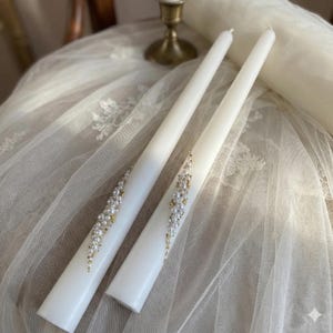 May include: Two white taper candles with pearl and gold bead embellishments. The candles are displayed on a white lace surface, with a brass candlestick in the background. The candles are a decorative item.
