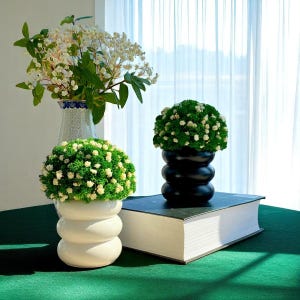 Puede incluir: Un bodegón con tres jarrones decorativos con arreglos florales. Un jarrón es blanco con un diseño apilado, otro es negro con un diseño apilado similar y el tercero es un jarrón alto con dibujos. Los jarrones están sobre una superficie verde.