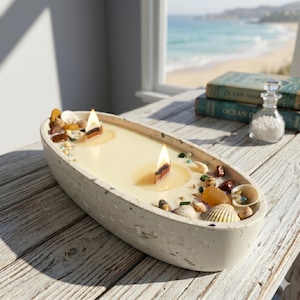 May include: An oval-shaped candle in a concrete container. The candle has two wooden wicks and is decorated with seashells and small stones. The candle sits on a wooden table with a view of the ocean in the background.