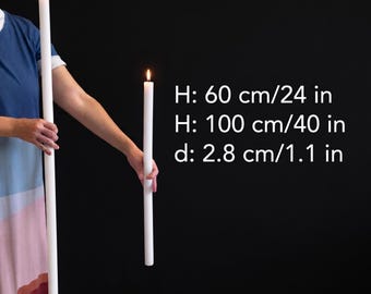 Puede incluir: Dos velas cónicas blancas, altas y delgadas, con llamas encendidas. El texto indica alturas de 60 cm y 100 cm, con un diámetro de 2,8 cm. Las velas son cilíndricas y lisas, proporcionando una fuente de luz brillante y constante.