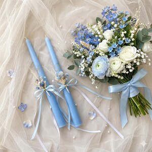 May include: Two pale blue candles adorned with dried flowers and ribbons, next to a bouquet of white and blue flowers. The bouquet is tied with a pale blue ribbon, creating a coordinated, elegant arrangement.