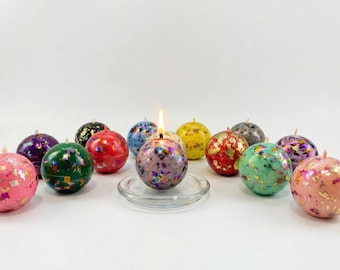 May include: A collection of colorful, spherical candles with metallic foil accents. One candle is lit, sitting on a clear glass dish. The candles are in various colors, including pink, green, red, and purple, with a white background.