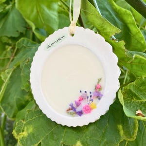 May include: A white, round ornament with a scalloped edge, featuring a clear center with colorful dried flowers. The ornament is suspended by a cream-colored ribbon. The text "OK.Decorstore" is printed on the ornament.