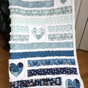 May include: A rag quilt featuring horizontal fabric strips in shades of blue, teal, and white. The quilt includes three heart-shaped patches with various patterns, including woodland animals, trees, and floral motifs, separated by frayed white seams.