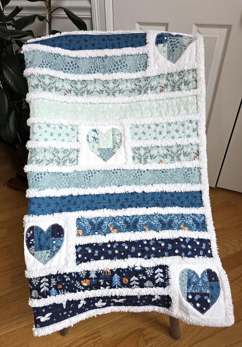 May include: A rag quilt featuring horizontal fabric strips in shades of blue, teal, and white. The quilt includes three heart-shaped patches with various patterns, including woodland animals, trees, and floral motifs, separated by frayed white seams.