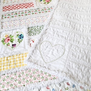 May include: A patchwork quilt featuring a variety of fabric strips with floral, gingham, and geometric patterns in red, green, yellow, and blue. The quilt includes white fabric sections with stitched heart outlines and frayed edges.