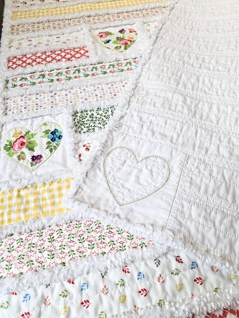 May include: A patchwork quilt featuring a variety of fabric strips with floral, gingham, and geometric patterns in red, green, yellow, and blue. The quilt includes white fabric sections with stitched heart outlines and frayed edges.