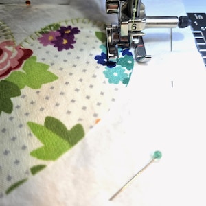 May include: A sewing machine presser foot stitching along the edge of a heart-shaped fabric applique. The fabric features a white background with a grey dot pattern, green leaves, and pink, purple, and blue flowers. A green-headed pin is visible.