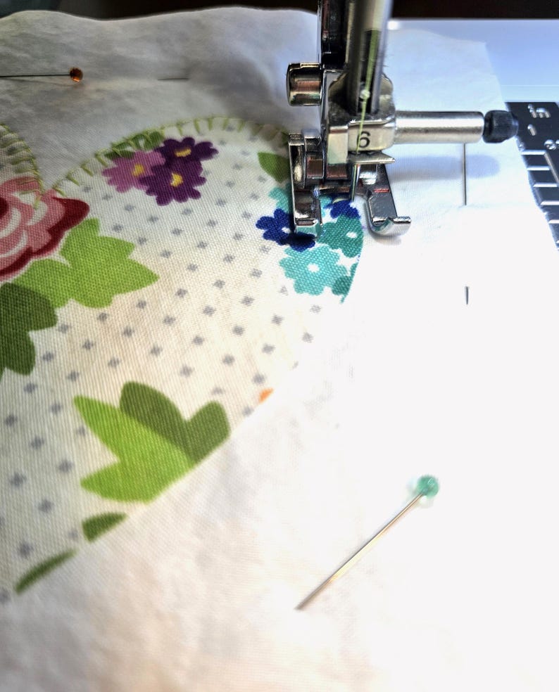 May include: A sewing machine presser foot stitching along the edge of a heart-shaped fabric applique. The fabric features a white background with a grey dot pattern, green leaves, and pink, purple, and blue flowers. A green-headed pin is visible.