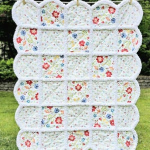 May include: A white floral quilt with a scalloped edge, hanging on a clothesline. The quilt features a repeating pattern of red, blue, and yellow flowers on a white background. The edges are trimmed with a fluffy white material.