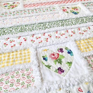 May include: A rag quilt featuring a patchwork of fabric squares with various floral, gingham, and leaf patterns in shades of pink, green, yellow, and red. The quilt includes several heart-shaped fabric appliqu&eacute;s and frayed white seams.