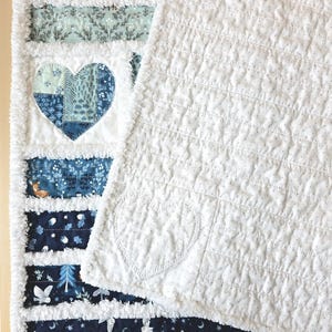 May include: A rag quilt featuring horizontal strips of fabric in shades of blue, white, and teal with patterns of owls, trees, and a deer. A large heart applique is centered on the left side, while the right side is a solid white fabric.
