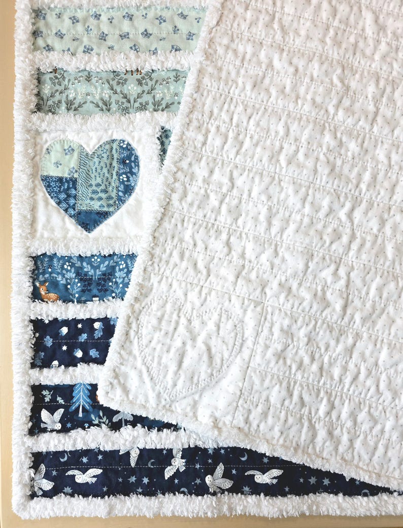 May include: A rag quilt featuring horizontal strips of fabric in shades of blue, white, and teal with patterns of owls, trees, and a deer. A large heart applique is centered on the left side, while the right side is a solid white fabric.