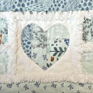 May include: A patchwork quilt featuring a central heart applique made of light green fabric with woodland animal and plant patterns. The heart is surrounded by white frayed fabric borders and a light blue background with tree and raccoon prints.