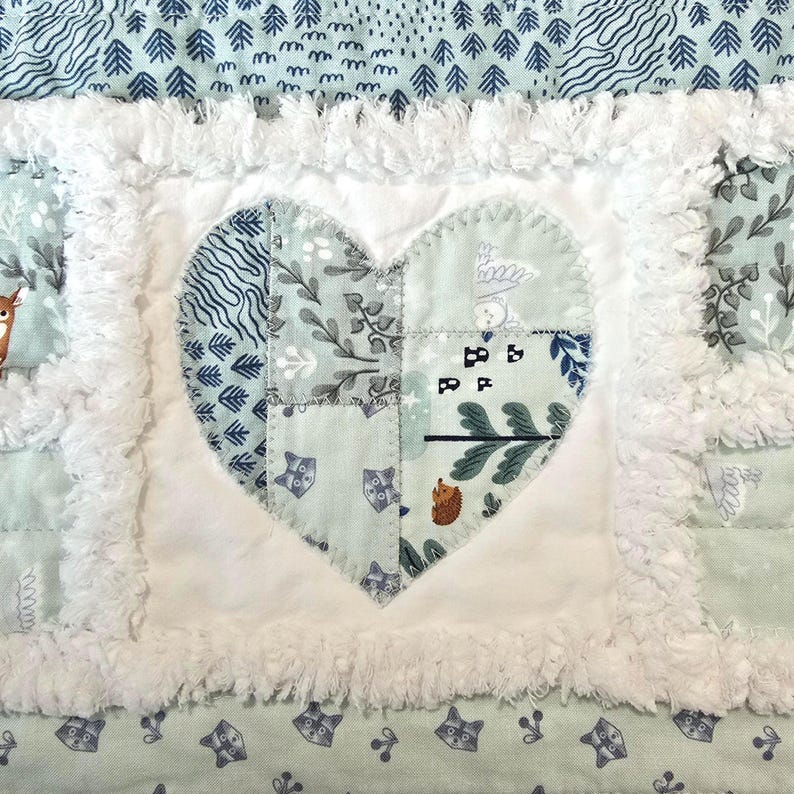 May include: A patchwork quilt featuring a central heart applique made of light green fabric with woodland animal and plant patterns. The heart is surrounded by white frayed fabric borders and a light blue background with tree and raccoon prints.