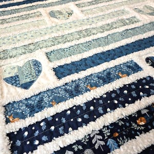 May include: A rag quilt featuring horizontal strips of fabric in shades of navy, royal blue, and light blue. The quilt includes patterns of deer, acorns, and trees, interspersed with white frayed seams and several heart-shaped fabric patches.