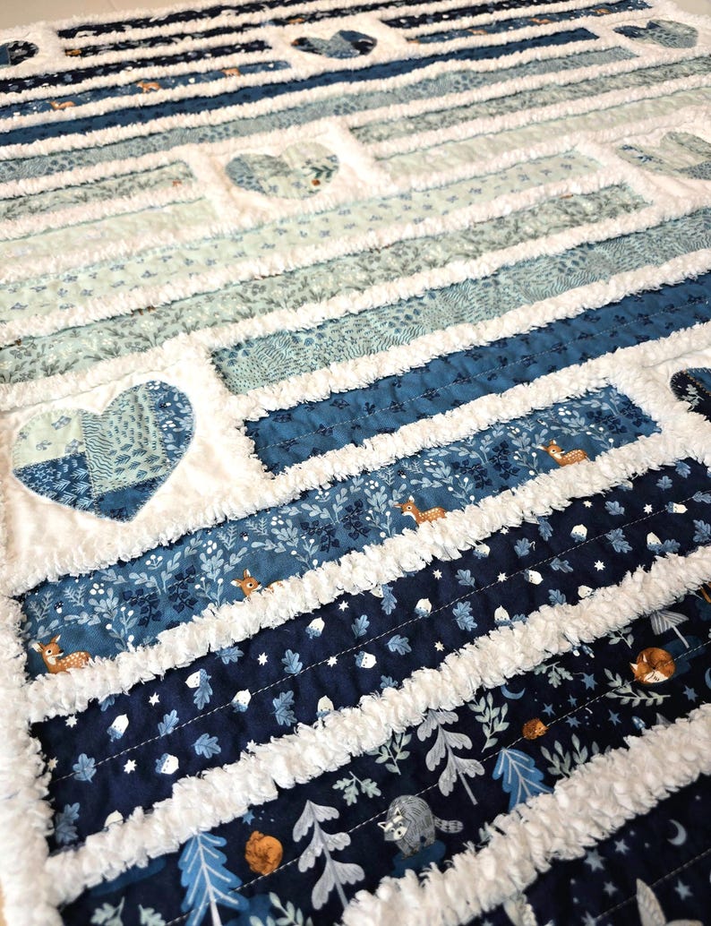 May include: A rag quilt featuring horizontal strips of fabric in shades of navy, royal blue, and light blue. The quilt includes patterns of deer, acorns, and trees, interspersed with white frayed seams and several heart-shaped fabric patches.