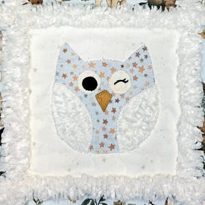 May include: A baby blanket featuring a central owl design. The owl is light blue with gold star accents, a black eye, and a winking eye. The owl is surrounded by white fluffy fabric and a patterned border with forest animals and foliage.