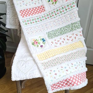 May include: A rag quilt featuring horizontal strips of white fabric with various floral, gingham, and geometric patterns in red, green, yellow, and blue. The quilt includes two fabric hearts with floral prints and one stitched heart outline.